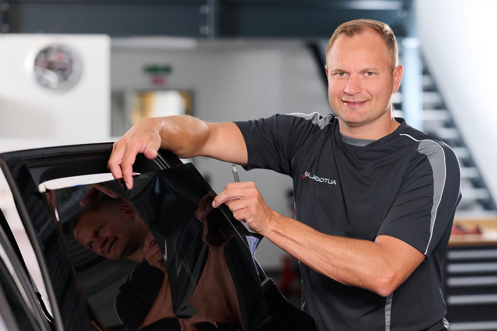 Eine Person in einem schwarzen Hemd bringt in einer Werkstatt mit einem Rakel eine Scheibentönungsfolie an einer Autoscheibe an.