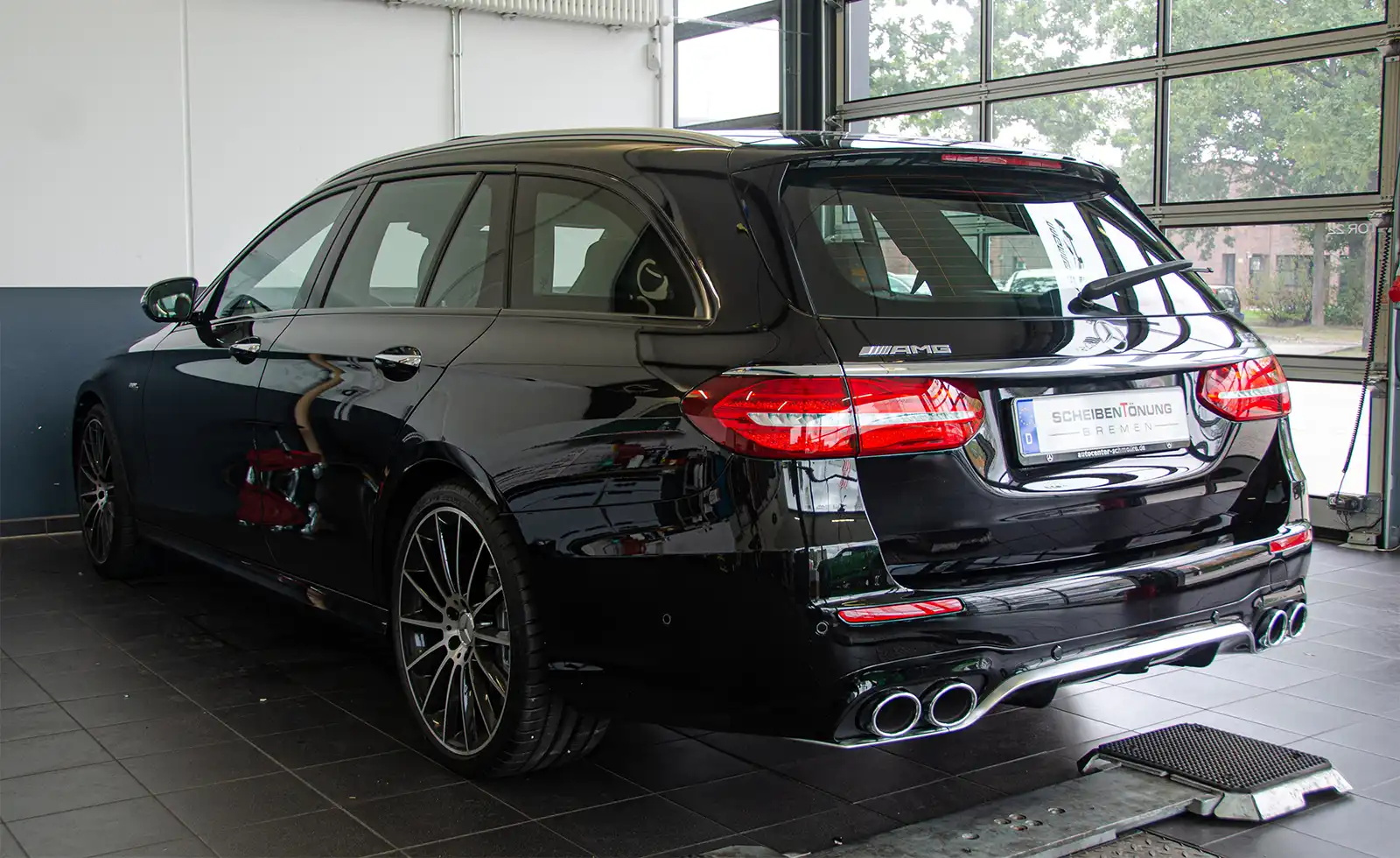 Ein schwarzer Mercedes-AMG-Kombi steht in einem Innenraum in der Nähe großer Fenster und präsentiert sich von hinten und von der Seite, wobei die AMG- und Modell-Embleme gut sichtbar sind.