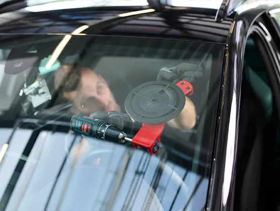 autoglas-austausch-scheibentoenung-bremen Eine Person verwendet ein Elektrowerkzeug und eine Absaugvorrichtung, um eine Windschutzscheibe von innen heraus zu reparieren oder auszutauschen.