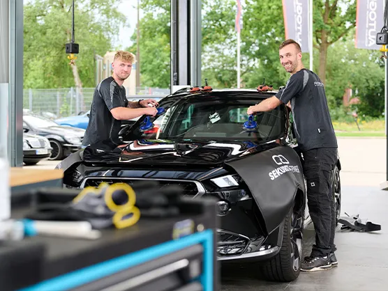 autoglas-austausch-scheibentoenung_bremen_ Zwei Männer montieren in einer Autowerkstatt mithilfe von Sauggreifern eine Windschutzscheibe an einem schwarzen Auto.