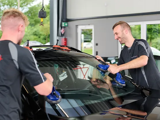 autoglas-austausch_scheibentoenung-bremen Zwei Männer montieren in einer Autowerkstatt mithilfe von Sauggreifern eine Windschutzscheibe an einem schwarzen Auto.